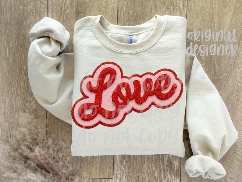 May include: Cream-colored sweatshirt with the word "Love" in a red, glittery, bubble letter design. The word is outlined in a lighter pink and red. The sleeves are rolled up.