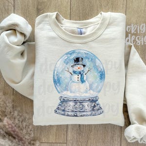 May include: Cream-colored sweatshirt featuring a watercolor illustration of a snowman inside a snow globe. The snowman wears a black top hat and a blue scarf. The snow globe sits on a decorative silver base. The words "original designer" are printed on the top right.