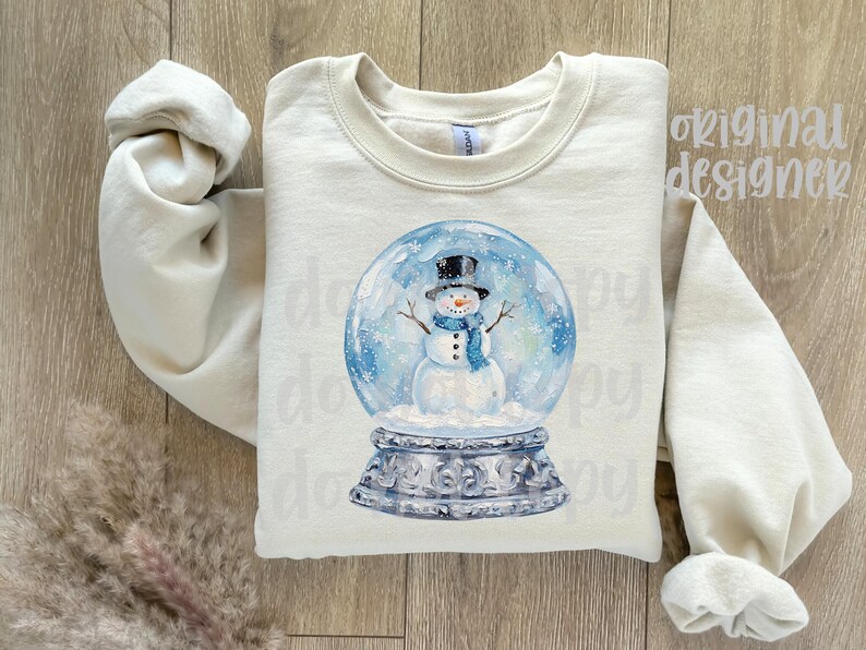 May include: Cream-colored sweatshirt featuring a watercolor illustration of a snowman inside a snow globe. The snowman wears a black top hat and a blue scarf. The snow globe sits on a decorative silver base. The words "original designer" are printed on the top right.