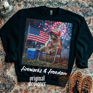 May include: Black sweatshirt featuring a raccoon wearing a red hat with "America" on it, holding a can with an American flag design, and sitting in a chair. The image includes a waving American flag, fireworks, and the text "fireworks & freedom original designer".