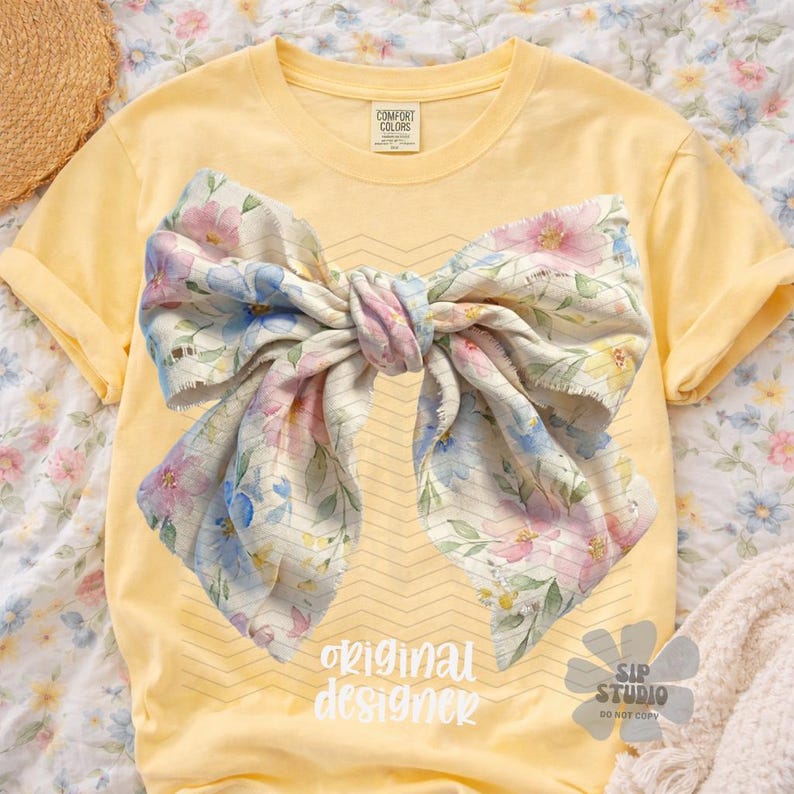 May include: A yellow t-shirt featuring a large bow design with a floral pattern in pink, blue, and yellow. The text "original designer" is printed in white. The shirt has rolled-up sleeves.