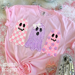 May include: A light pink t-shirt featuring three ghost graphics. The left ghost has a pink and white checkerboard pattern, the middle ghost is purple with a white bow, and the right ghost is pink with small purple and white flower accents.