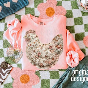 May include: A light pink sweatshirt featuring a floral chicken design. The chicken is made of a floral fabric with a quilted appearance. The words "original designer" are printed in a gothic font on the bottom right of the image.