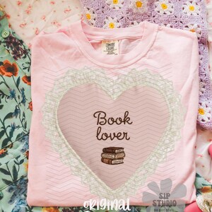 May include: A pale pink t-shirt featuring a heart design with the words "Book lover" and a stack of books. The heart is framed by a lace-like border. The shirt rests on a floral patterned surface.