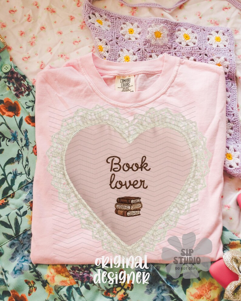 May include: A pale pink t-shirt featuring a heart design with the words "Book lover" and a stack of books. The heart is framed by a lace-like border. The shirt rests on a floral patterned surface.
