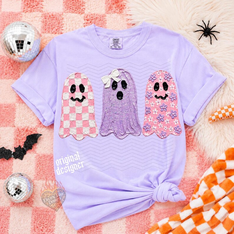 May include: A light purple short-sleeve t-shirt featuring three ghost illustrations. The left ghost has a pink and white checkered pattern, the middle ghost is purple with a white bow, and the right ghost is pink with small purple flowers.