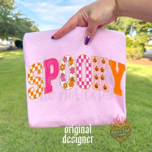 May include: A light pink t-shirt featuring the word SPOOKY in large, decorative letters. The letters have varied patterns including orange and white checkers, solid hot pink, floral and ghost motifs, pink checkers, and orange pumpkins.