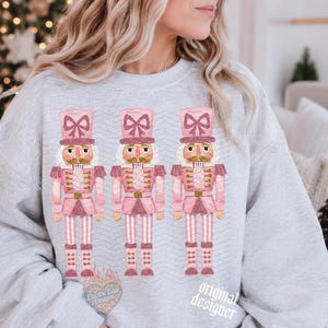 May include: A light gray crewneck sweatshirt featuring a graphic print of three identical nutcracker figures. Each nutcracker is colored in shades of pink and white with glittery accents, wearing a tall hat with a bow, and standing in a row.