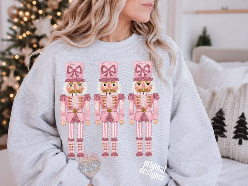 May include: A light gray crewneck sweatshirt featuring a graphic print of three identical nutcracker figures. Each nutcracker is colored in shades of pink and white with glittery accents, wearing a tall hat with a bow, and standing in a row.