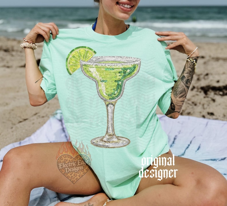May include: A light green t-shirt featuring a sequined margarita glass design with a lime wedge. The shirt has short sleeves and a relaxed fit. The words "original designer" are printed on the shirt.
