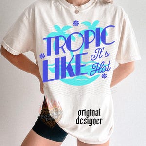 May include: An off-white short-sleeve t-shirt featuring a graphic print with the text TROPIC LIKE It's Hot in blue lettering. The design includes light blue palm tree silhouettes, small blue flower icons, and wavy line patterns across the chest area.