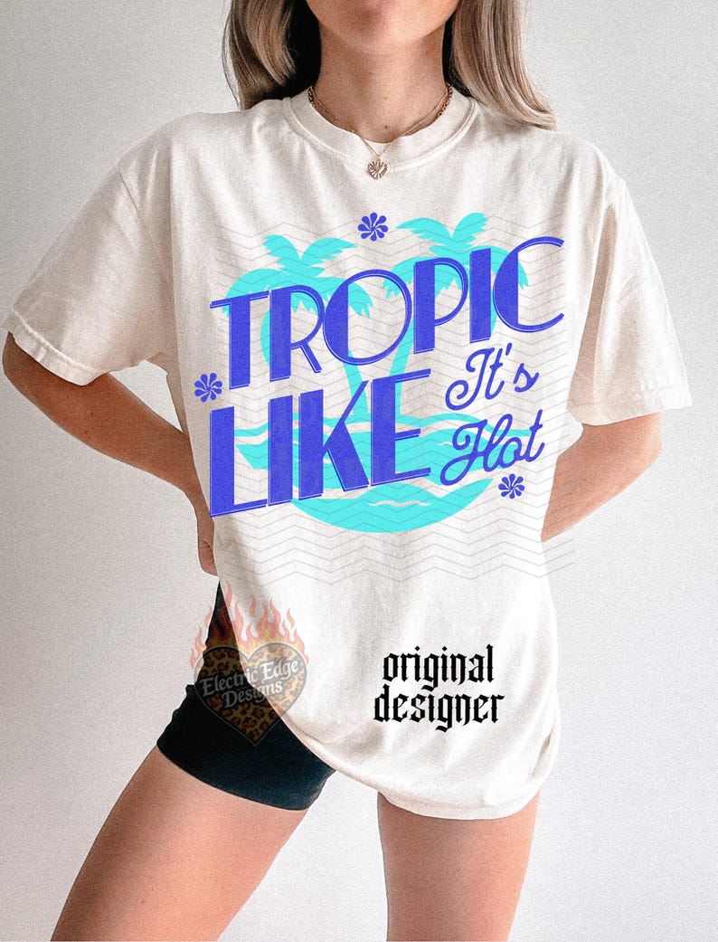 May include: An off-white short-sleeve t-shirt featuring a graphic print with the text TROPIC LIKE It's Hot in blue lettering. The design includes light blue palm tree silhouettes, small blue flower icons, and wavy line patterns across the chest area.