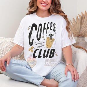 May include: White t-shirt with the words "COFFEE CLUB" in a bold, black font, with the phrase "running on caffeine" below. A graphic of a coffee cup and coffee beans are also featured. The shirt is a casual, everyday wear item.