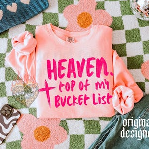 May include: A light pink sweatshirt with the words "HEAVEN, top of my BUCKET LIST" in bright pink. The sweatshirt is on a green and white checkered surface with a pink flower rug and a pair of blue jeans.