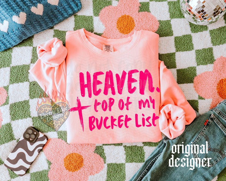 May include: A light pink sweatshirt with the words "HEAVEN, top of my BUCKET LIST" in bright pink. The sweatshirt is on a green and white checkered surface with a pink flower rug and a pair of blue jeans.