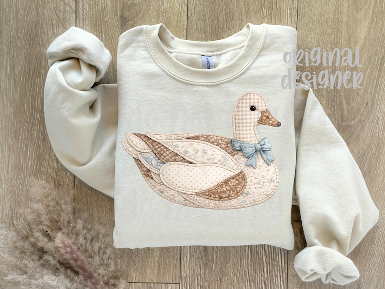 May include: Cream-colored sweatshirt featuring a patchwork duck design with a checkered head, brown and beige wings, and a light blue bow. The sleeves are rolled up, and the text "original designer" is visible.