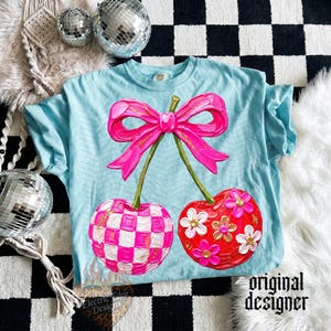 May include: A light blue t-shirt featuring a large graphic of two cherries connected by a bright pink bow. One cherry has a pink and white checkered pattern, while the other is solid red with white and gold glitter flowers on the surface.
