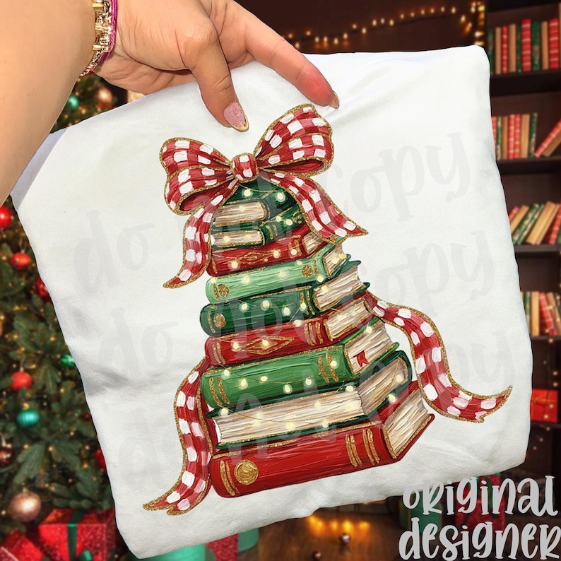 May include: A white t-shirt featuring a graphic illustration of a stack of books arranged in a triangular tree shape. The stack is topped with a large red and white checkered bow and decorated with small yellow lights, with a matching ribbon draped.