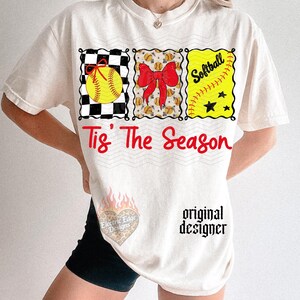 May include: An off-white t-shirt featuring three rectangular graphics with a softball, a red bow, and the word Softball. Below, the red text reads Tis' The Season. A small heart-shaped logo with flames and the text Electric Edge Designs is visible.