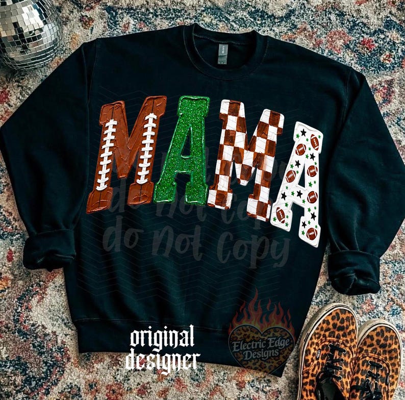 May include: A black long-sleeve sweatshirt featuring the word MAMA in large letters. The M is brown with white football laces, the A is green glitter, the M is a brown and white checkerboard pattern, and the A is white with footballs and stars.