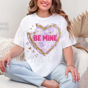 Op de afbeelding: Wit T-shirt met een paars hartontwerp, gouden accenten en de roze tekst "BE MINE". Er is een hap uit het hart genomen. Het T-shirt wordt door een persoon gedragen.