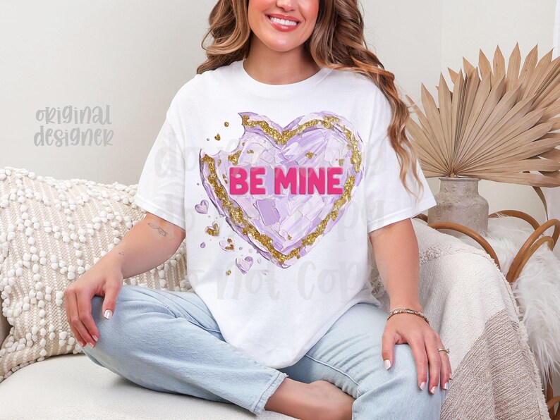 Op de afbeelding: Wit T-shirt met een paars hartontwerp, gouden accenten en de roze tekst "BE MINE". Er is een hap uit het hart genomen. Het T-shirt wordt door een persoon gedragen.