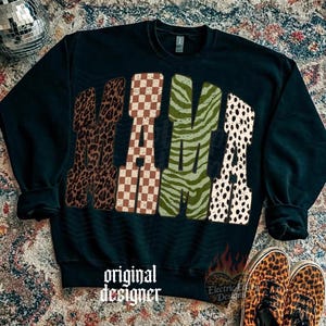 May include: A black long-sleeve sweatshirt featuring the word MAMA in large block letters. Each letter has a distinct pattern: the first M is brown leopard print, the A is a brown and white checkerboard, the second M is green zebra print, and the final A is a white and black dalmatian spot print. The text original designer is printed in white below.
