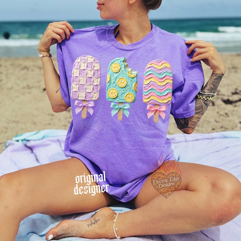 May include: A purple short-sleeve t-shirt featuring a graphic of three popsicles with textured, raised-style details. The left popsicle has a checkered pattern, the center has smiley faces, and the right has wavy lines, each with a small bow.