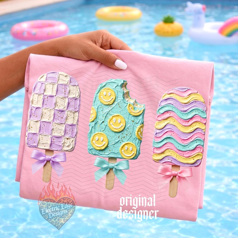 May include: A light pink t-shirt featuring three popsicle graphics with textured frosting. The left popsicle has a purple and white checkerboard pattern, the middle is light blue with yellow smiley faces, and the right has wavy stripes.