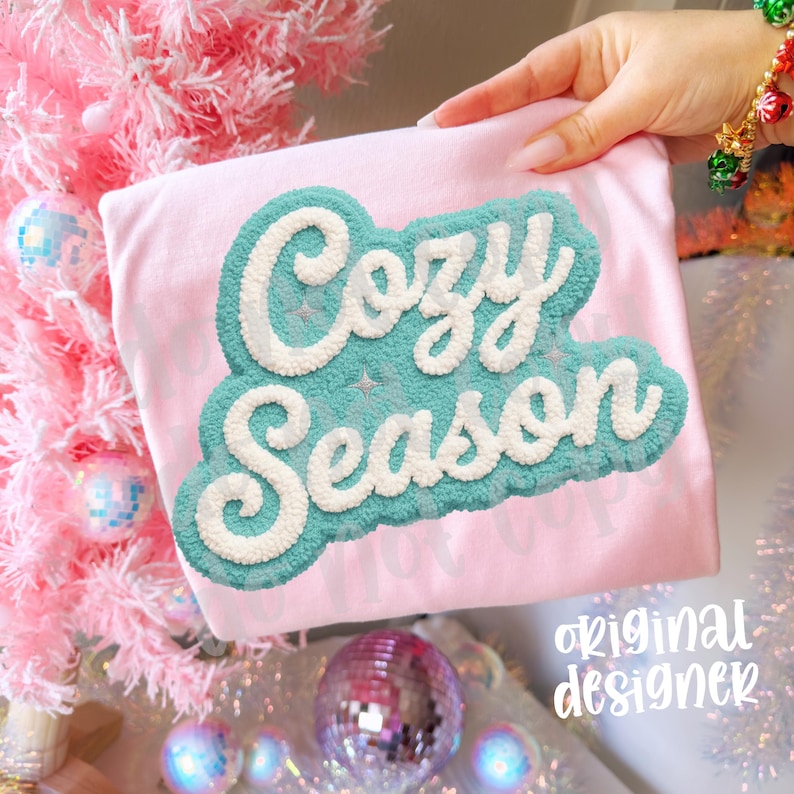 May include: A light pink t-shirt with the words "Cozy Season" in a teal and white textured design. The shirt is held up in front of a pink Christmas tree with disco ball ornaments.