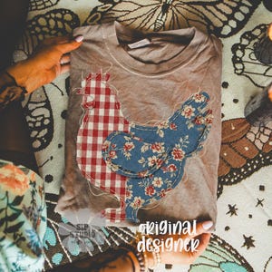 May include: A tan crew neck t-shirt featuring a patchwork chicken design. One side of the chicken is red and white gingham, the other is blue floral. The words "original designer" are visible in the image.