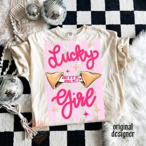 May include: A cream-coloured t-shirt with a pink graphic. The graphic features the words "Lucky Girl" in a cursive font, a fortune cookie, and the text "What if it all works out?" The t-shirt has a relaxed fit.