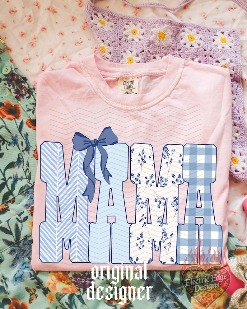以下が含まれることがあります： 大きな文字で「MAMA」と書かれた、ピンク色のComfort Colors Tシャツ。文字は青い模様と青いリボンで装飾されています。「original designer」の文字が下にあります。