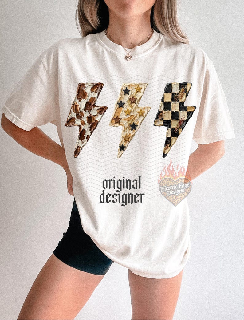 May include: Cream-colored t-shirt featuring three lightning bolt designs with unique patterns: cow print, stars, and a black and tan checkerboard. The shirt has the text "original designer" in a gothic font.