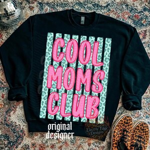Peut inclure: Sweat-shirt noir avec l'inscription "COOL MOMS CLUB" en rose, soulign&eacute;e d'un rose plus fonc&eacute;. Le texte est plac&eacute; sur un fond bleu clair avec des rayures verticales et un motif l&eacute;opard. Les mots "original designer" sont en bas.