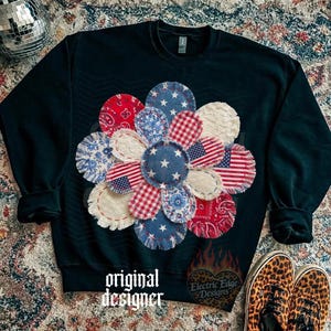 May include: A dark green sweatshirt featuring a large flower design made of fabric patches in red, white, and blue patterns. The flower includes stars, stripes, and American flag motifs. The text "original designer" is at the bottom.