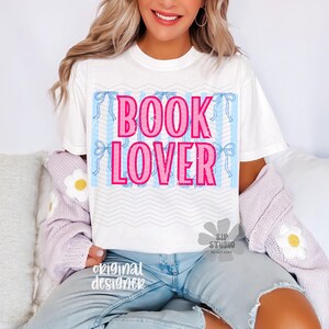May include: White t-shirt with the text "BOOK LOVER" in pink, set against a light blue striped background with pink bows. The shirt is part of a casual outfit.