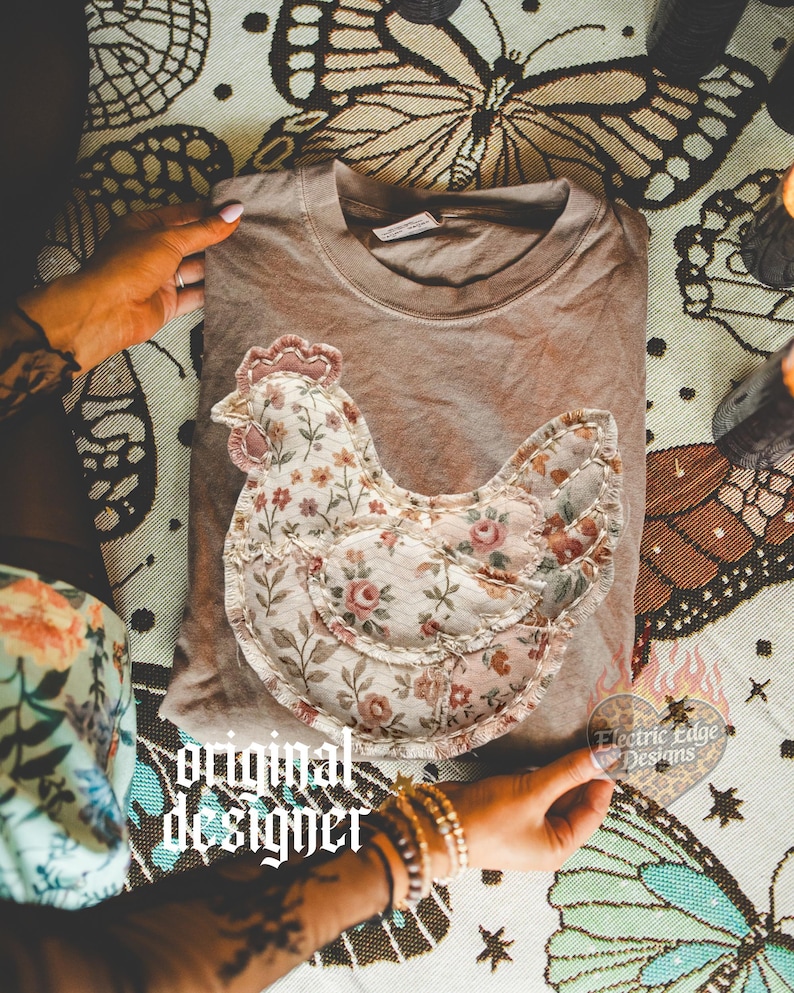 May include: A brown t-shirt with a floral chicken applique. The chicken design features a mix of floral patterns in pink, cream, and brown. The words "original designer" are printed in a gothic font. A heart-shaped sticker with the text "Electric Edge Designs" is also visible.
