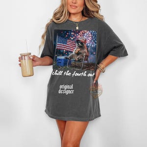 K&ouml;nnte beinhalten: Dunkelgraues T-Shirt mit einem Aufdruck eines Waschb&auml;ren mit Hut, der in einem Stuhl sitzt, mit einer amerikanischen Flagge im Hintergrund. Der Text "chill the fourth out" und "original designer" ist auf dem Shirt aufgedruckt.