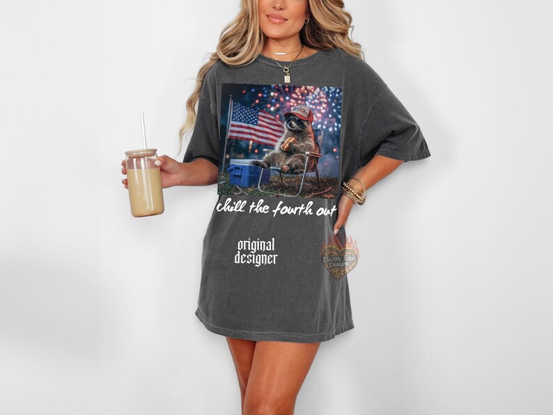 K&ouml;nnte beinhalten: Dunkelgraues T-Shirt mit einem Aufdruck eines Waschb&auml;ren mit Hut, der in einem Stuhl sitzt, mit einer amerikanischen Flagge im Hintergrund. Der Text "chill the fourth out" und "original designer" ist auf dem Shirt aufgedruckt.