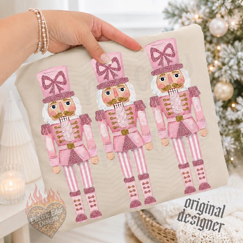 May include: A beige fabric item featuring a repeating illustration of three pink nutcrackers. Each nutcracker wears a tall hat with a glittery pink bow, a pink uniform with gold accents, and striped pants. The text Electric Edge Designs is visible.