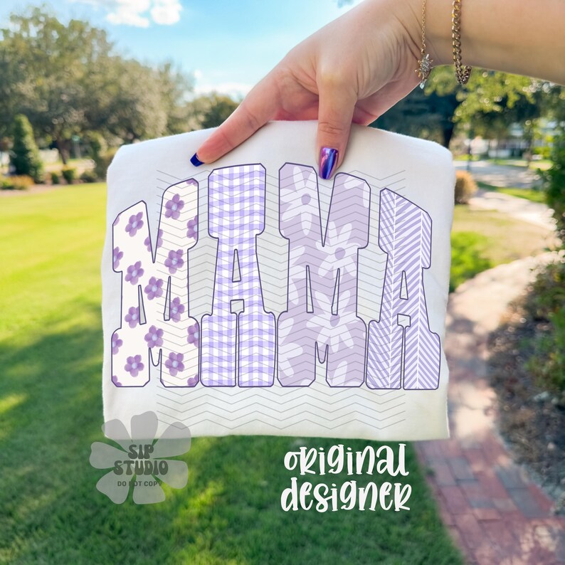 May include: White t-shirt with the word "MAMA" in large, lavender letters. The letters have different patterns, including floral and gingham. The shirt is held up against a background of green grass and trees.