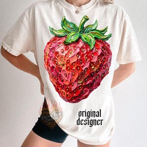 May include: Cream-colored t-shirt featuring a large, detailed strawberry illustration. The strawberry is rendered in shades of red, pink, and gold, with green leaves. The text "original designer" is printed in black. The shirt has short sleeves.
