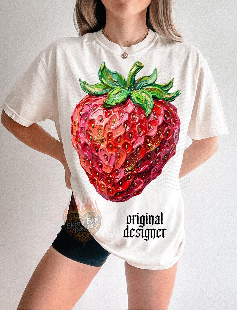 May include: Cream-colored t-shirt featuring a large, detailed strawberry illustration. The strawberry is rendered in shades of red, pink, and gold, with green leaves. The text "original designer" is printed in black. The shirt has short sleeves.