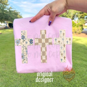 May include: A light pink t-shirt featuring three vertical cross designs. The left cross has a blue floral pattern, the center cross has a beige and white checkerboard pattern, and the right cross has a pattern of small blue and brown crosses.