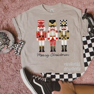 Peut inclure: T-shirt beige avec trois soldats casse-noisettes de style aquarelle en rouge, blanc et or. Les mots "Merry Christmas" sont imprim&eacute;s en dessous. Le t-shirt porte &eacute;galement les mots "original designer".