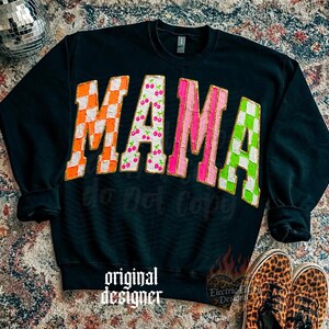 May include: Black sweatshirt with the word "MAMA" in large, colorful letters. The letters have orange checkerboard, cherry print, pink stripes, and green checkerboard patterns. The words "do not copy" and "original designer" are also visible.