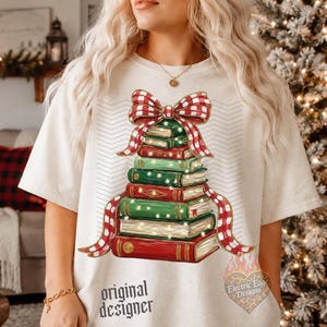May include: A cream-colored t-shirt featuring a graphic of a stack of books arranged in a tree shape, topped with a red and white checkered bow. The books are green and red with gold accents and small lights. The text original designer is printed below.