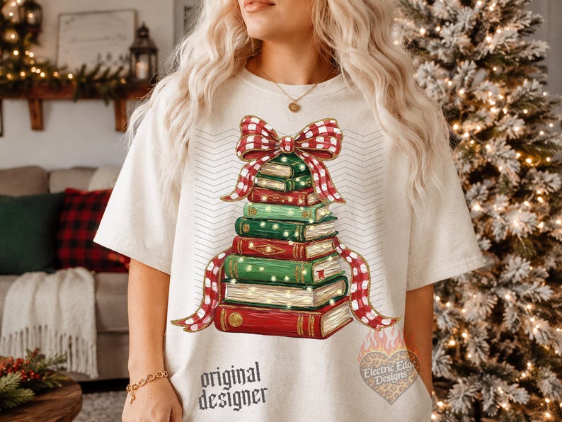 May include: A cream-colored t-shirt featuring a graphic of a stack of books arranged in a tree shape, topped with a red and white checkered bow. The books are green and red with gold accents and small lights. The text original designer is printed below.