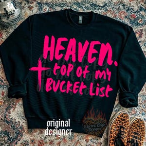 May include: Black sweatshirt with the words "HEAVEN. top of my BUCKET List" in bright pink. A pink cross is on the left side. The words "original designer" are at the bottom left, and a logo is on the bottom right.